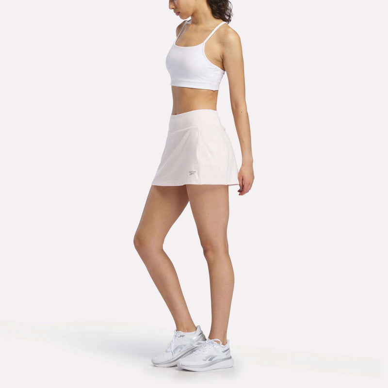 A woman in a white sports bra, Reebok Women's Break Point Skort with built-in compression shorts, and white sneakers stands against a plain white background, facing sideways with her arms relaxed at her sides.