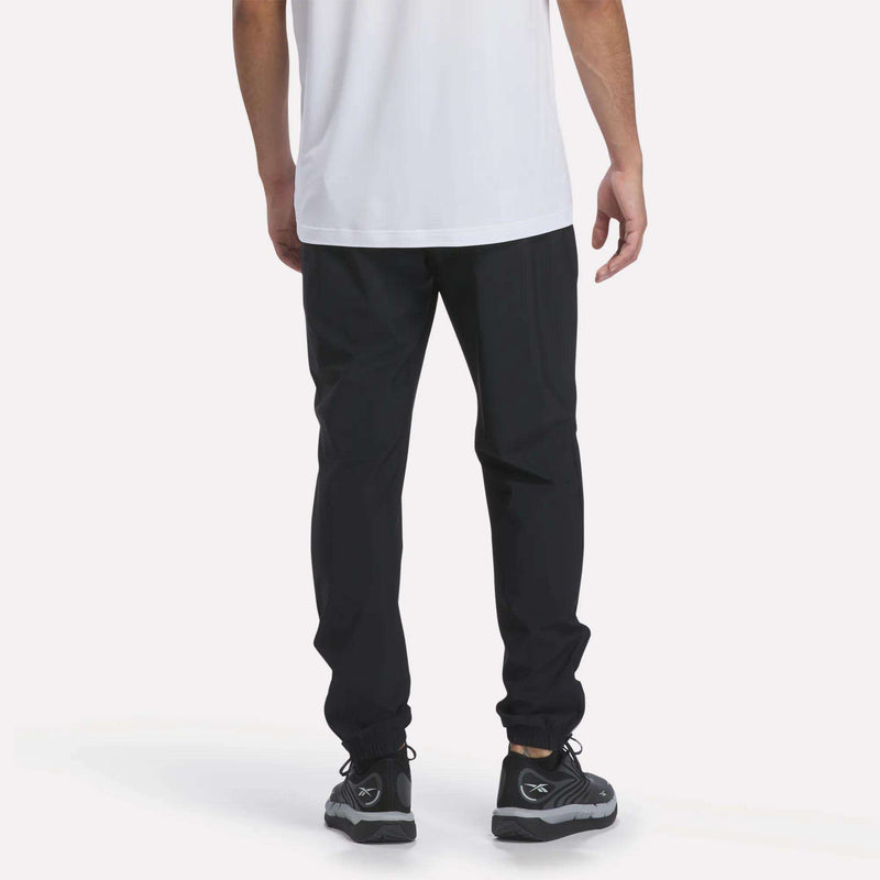 A person wearing a white shirt, Reebok Men’s Repeat Pants in a slim fit, and black sneakers is standing with their back facing the camera against a plain light background.