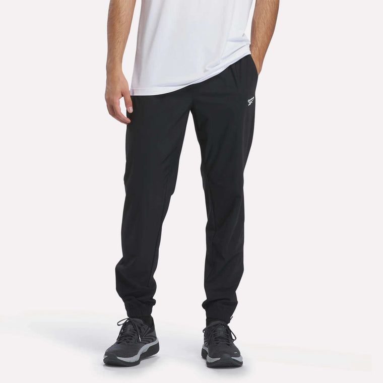 A person wearing a white shirt, black Reebok Men’s Repeat Pants, and black sneakers stands with one hand in their pocket against a plain white background.