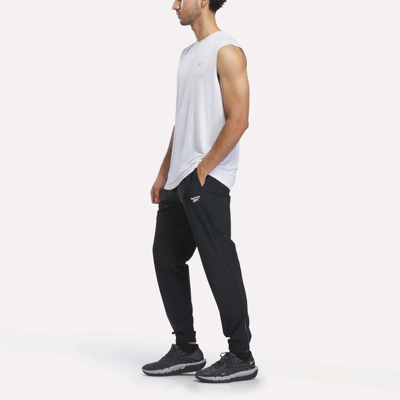 A man wearing a sleeveless white shirt, slim fit black Men’s Repeat Pants by Reebok, and black sneakers stands with his hands in his pockets against a plain white background. The Reebok Men’s Repeat Pants feature a cooling design for all-day comfort.