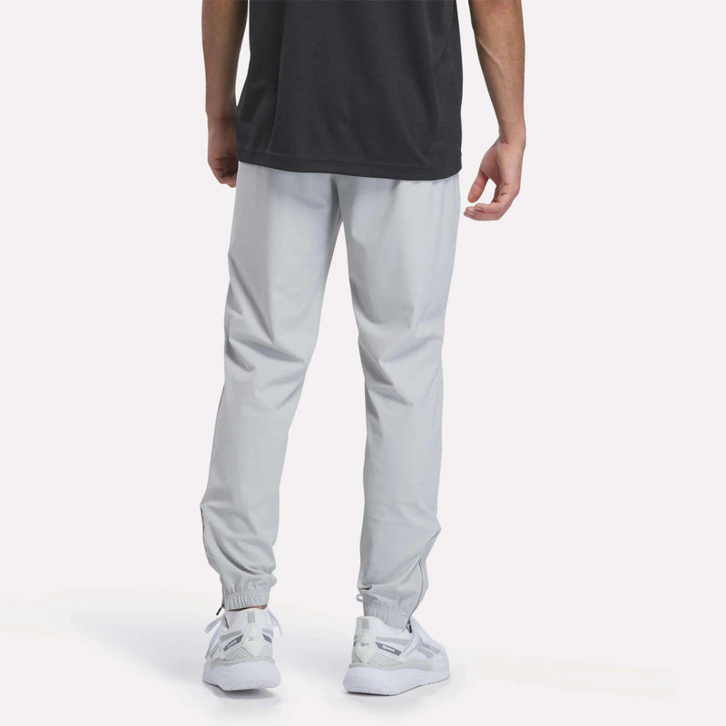 A person is shown from the back against a plain white background, wearing Reebok Men’s Repeat Pants in light gray, paired with a black shirt and white sneakers.