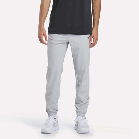 A person stands against a plain, light background wearing Men’s Repeat Pants by Reebok in Grey 1, paired with a black shirt and white sneakers. Only the lower half of the body is visible, highlighting the slim fit joggers’ tapered design.