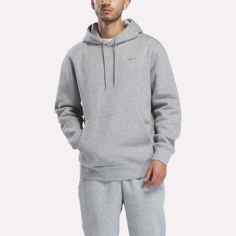 A person wearing the Reebok Men’s Patent Hoodie in Grey Heather, featuring an embroidered logo, and matching sweatpants stands with hands in the hoodie pocket against a plain white background.