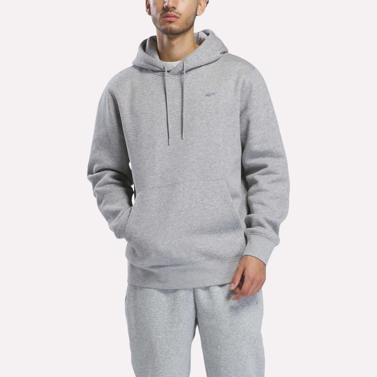 A person wearing the Reebok Men’s Patent Hoodie in Grey Heather, featuring an embroidered logo, and matching sweatpants stands with hands in the hoodie pocket against a plain white background.