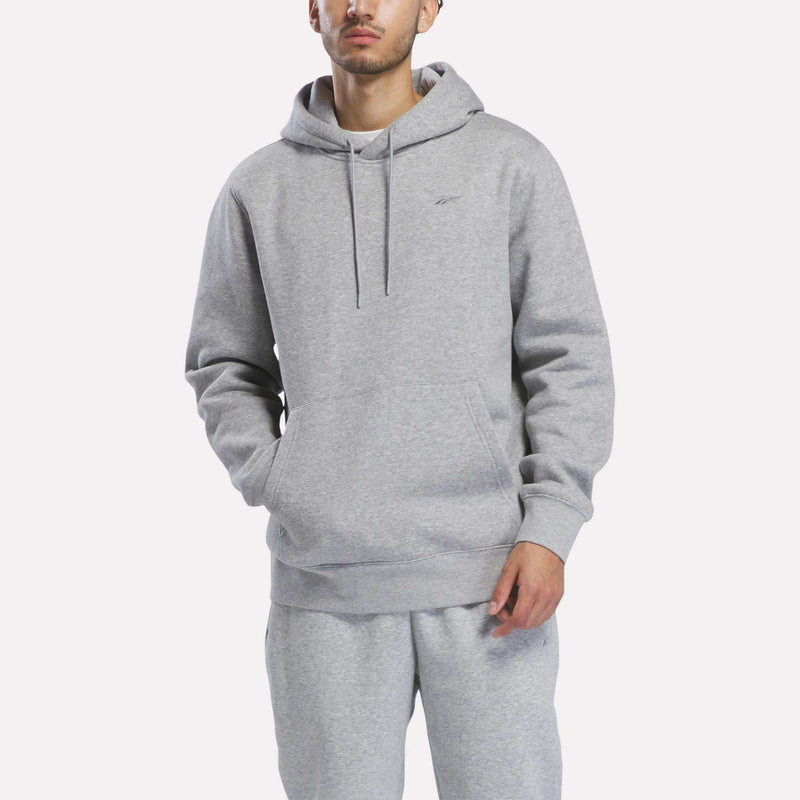 A person wearing the Reebok Men’s Patent Hoodie in Grey Heather, featuring an embroidered logo, and matching sweatpants stands with hands in the hoodie pocket against a plain white background.