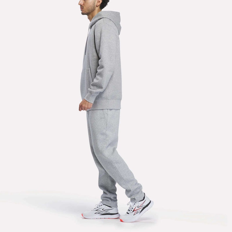 A person stands in profile on a plain white background, wearing the Reebok Men’s Patent Hoodie in gray paired with matching sweatpants and white athletic shoes featuring black and red accents. The hoodie is finished with an embroidered logo, offering a subtle touch of style to the overall look.