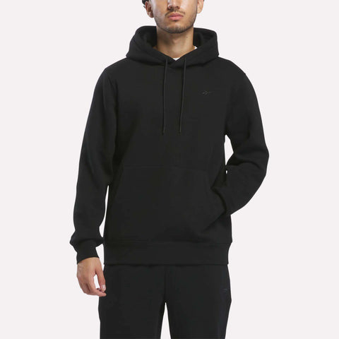 A person wearing the Men’s Patent Hoodie by Reebok in black and black pants stands against a white background, with one hand in their pocket and the other hanging by their side.