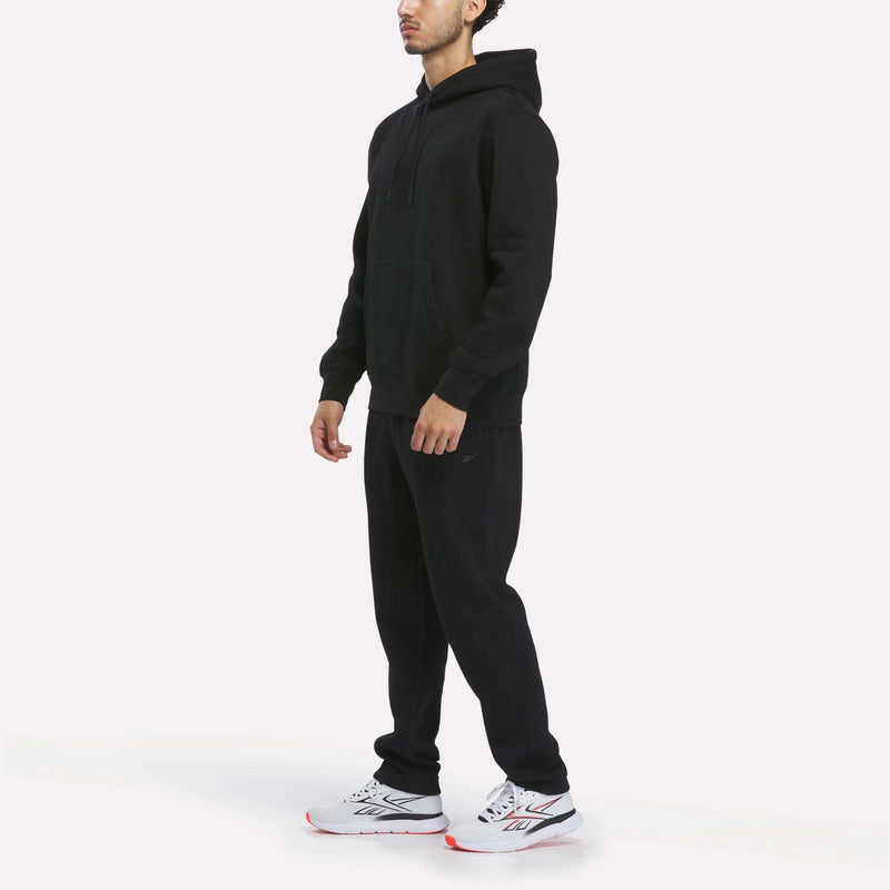 A person standing and wearing the Reebok Men’s Patent Hoodie in black, paired with matching black sweatpants and white athletic sneakers with red accents, on a plain white background.