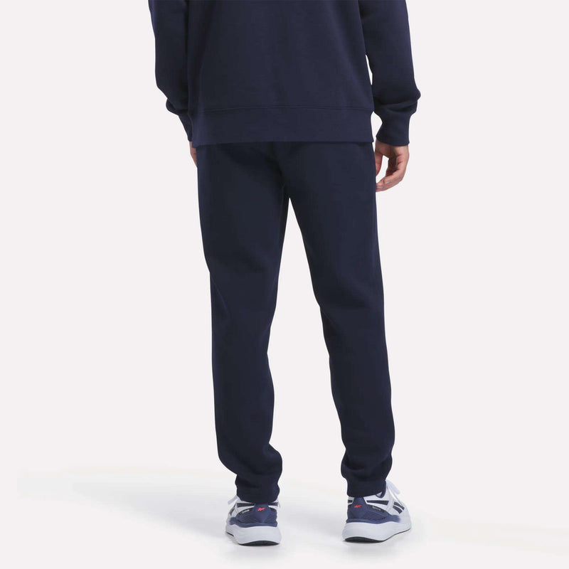 A person stands facing away on a plain light background, showing only the lower half of their body. They are wearing Reebok Men’s Patent Pant and a matching navy blue sweatshirt made from soft knit fabric, featuring an elastic waistband, paired with white and blue athletic shoes.