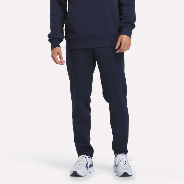 A person is shown from the waist down wearing Reebok Men’s Patent Pant in Vector Navy, featuring a matching navy color and elastic waistband, crafted from soft knit fabric. The outfit is paired with white athletic shoes and photographed against a plain white background.
