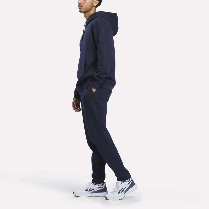 A person stands sideways wearing a navy blue hoodie, Reebok Men’s Patent Pant made from soft knit fabric with an elastic waistband, and white sneakers against a plain white background.