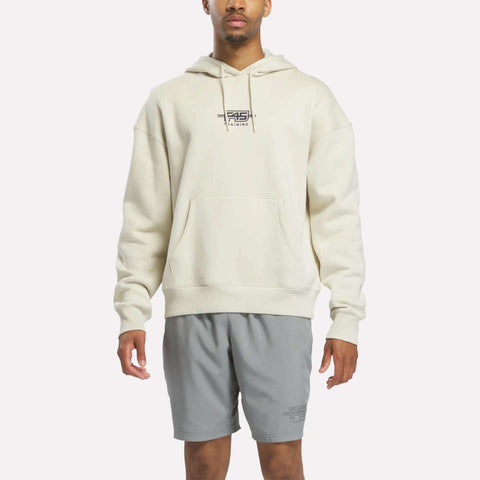 A person wearing the Reebok x F45 Training Relaxed Fleece Hoodie in Linen—a cream-colored, relaxed-fit hoodie featuring "F45 Training" on the chest—paired with light gray athletic shorts, stands against a plain white background.