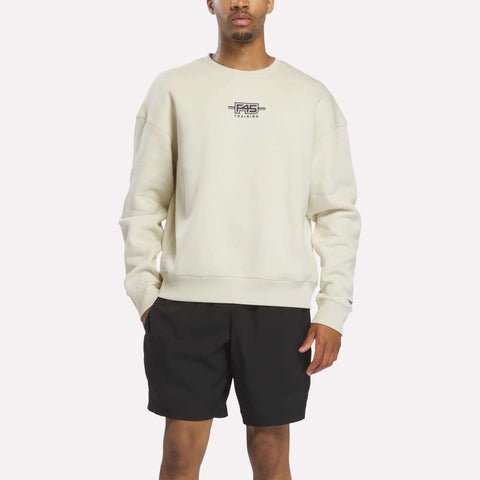 A person stands against a plain white background wearing the Reebok x F45 Training Relaxed Graphic Sweatshirt in Linen with a small chest logo, paired with black shorts.