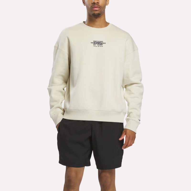 A person stands against a plain white background wearing the Reebok x F45 Training Relaxed Graphic Sweatshirt in Linen with a small chest logo, paired with black shorts.