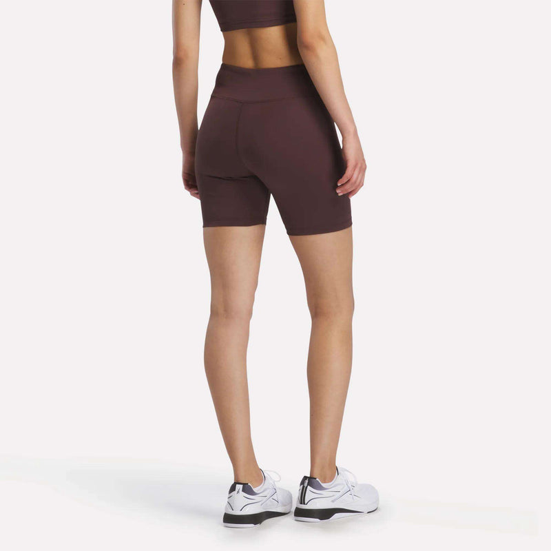 A person stands facing away, displaying the back view while wearing the Women's Reebok x F45 Training High-Rise Biker Shorts in burgundy with a matching sports bra. They are also wearing white Reebok athletic shoes, and the background is plain white.