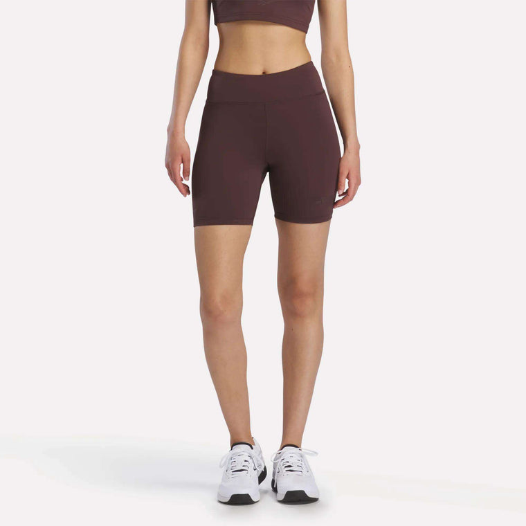 A woman stands facing forward against a plain white background, wearing Reebok x F45 Training High-Rise Biker Shorts in burgundy with a matching sports bra and white Reebok sneakers.