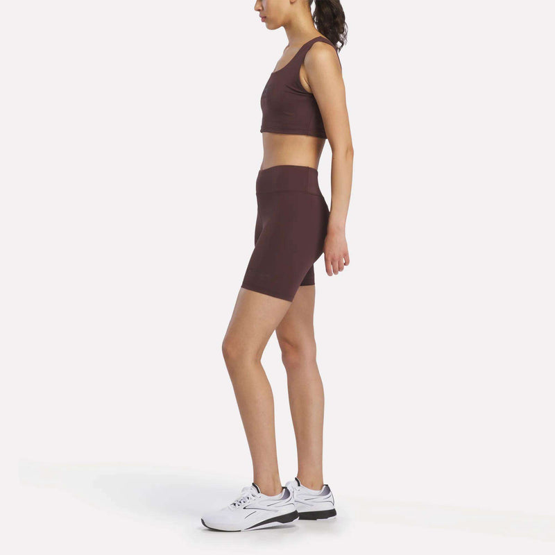 A woman stands in profile against a plain white background, wearing maroon Women's Reebok x F45 Training High-Rise Biker Shorts and a matching sports bra with white athletic shoes, ready for her next F45 Training session.