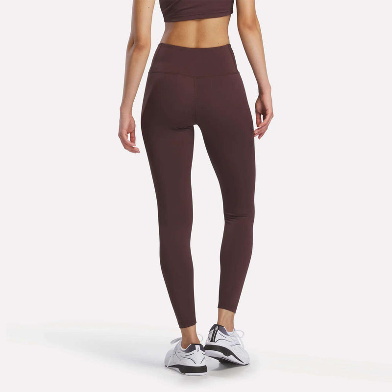A woman stands facing away, showcasing the back view of her outfit: she wears a matching burgundy sports bra and the Women's Reebok x F45 Training Leggings, paired with white Reebok performance athletic shoes.