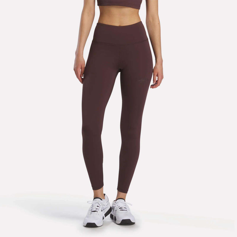 Wearing the Women’s Reebok x F45 Training Leggings and a maroon sports bra, a woman stands with her arms relaxed, paired with white Reebok performance athletic shoes against a plain background.