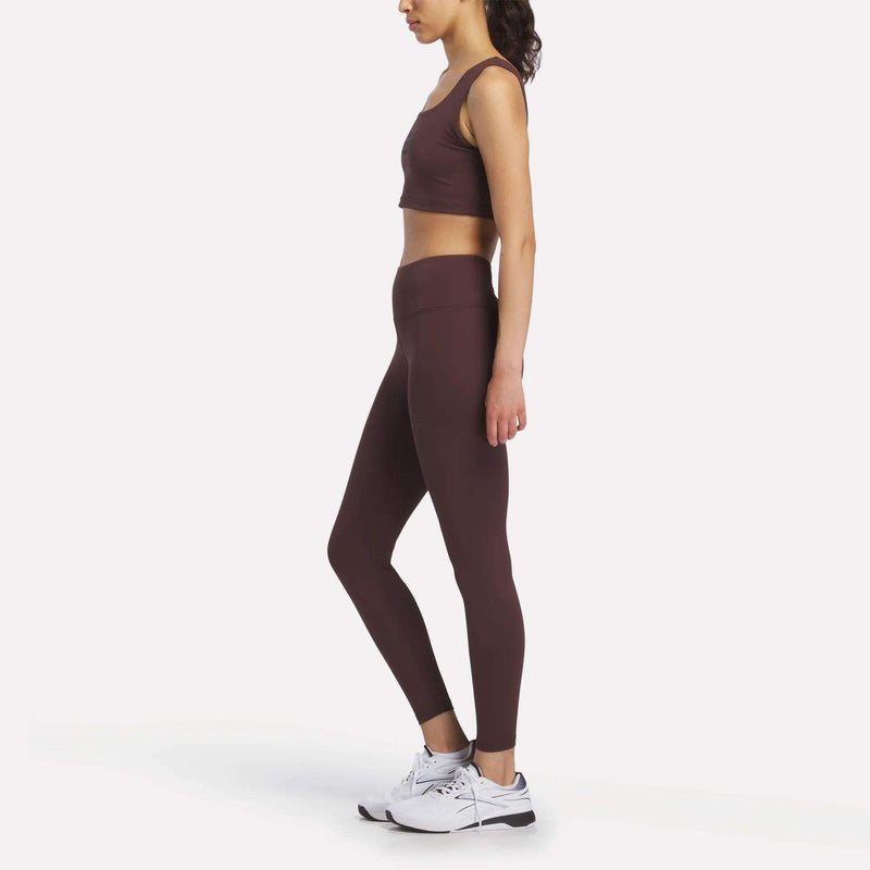 A woman stands in profile against a plain white background, wearing Women's Reebok x F45 Training Leggings and a burgundy sports bra, paired with white Reebok athletic shoes.