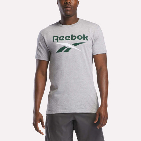 A man wears a Reebok Men's Stacked Vector Graphic T-Shirt in light gray, 100% cotton, with a large green Reebok logo and dark gray shorts, standing against a plain light background.