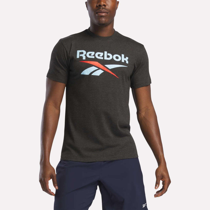 A man in a dark gray Reebok Men's Stacked Vector Graphic T-Shirt, 100% cotton with a bold white and red chest logo, paired with navy blue shorts, stands against a plain light background.