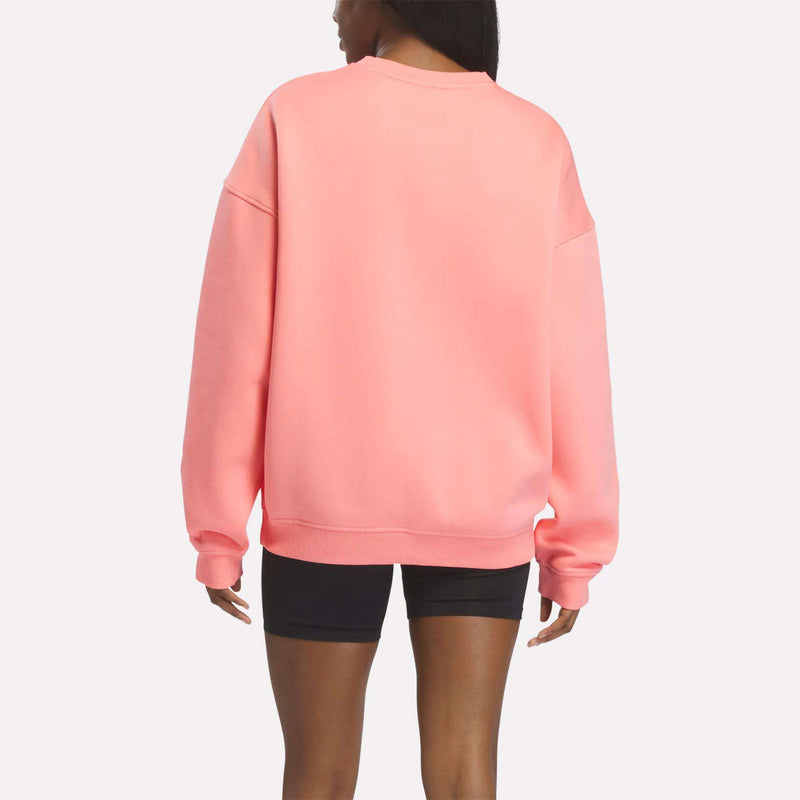 A person is shown from the back standing against a plain white background, wearing a relaxed-fit Reebok Crewneck Sweatshirt in pink paired with black shorts, highlighting a modern heritage style.