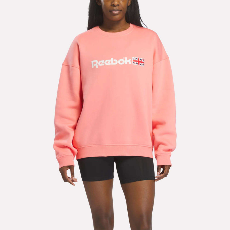 A person wearing a relaxed-fit Reebok Crewneck Sweatshirt in Canndy Pink, paired with black shorts and featuring the UK flag logo, stands against a plain white background, effortlessly showcasing classic heritage style.