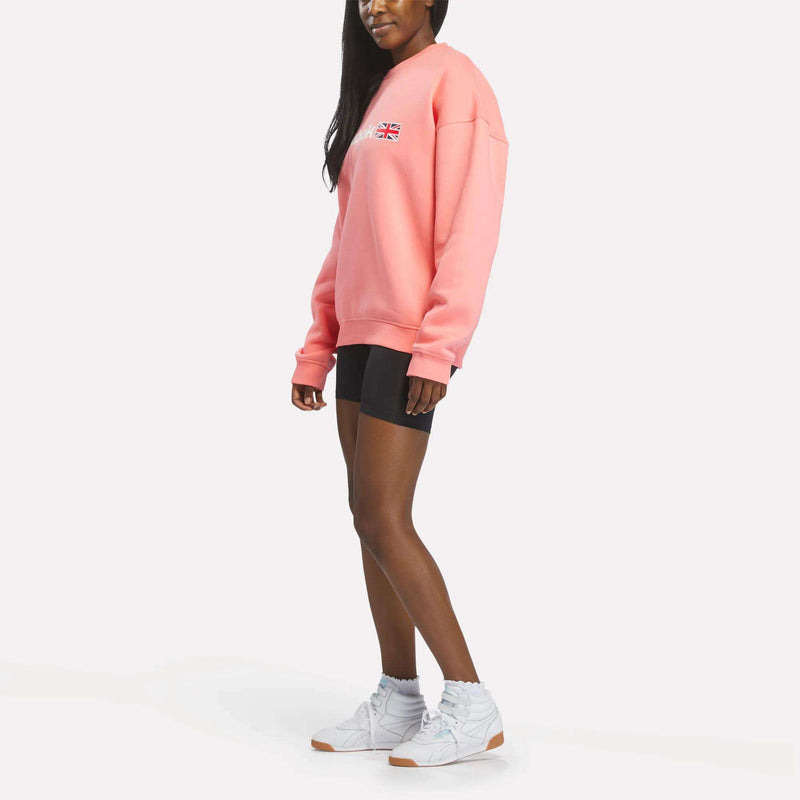 A woman wearing a relaxed fit pink Reebok Crewneck Sweatshirt, black shorts, and white sneakers stands against a white background, facing slightly to the side.