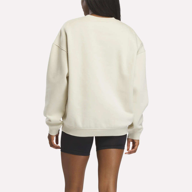 A person stands facing away from the camera against a plain white background, wearing a loose-fitting, cream-colored Reebok Crewneck Sweatshirt with black shorts. The classic Union Jack logo is subtly featured on the back of the sweatshirt.