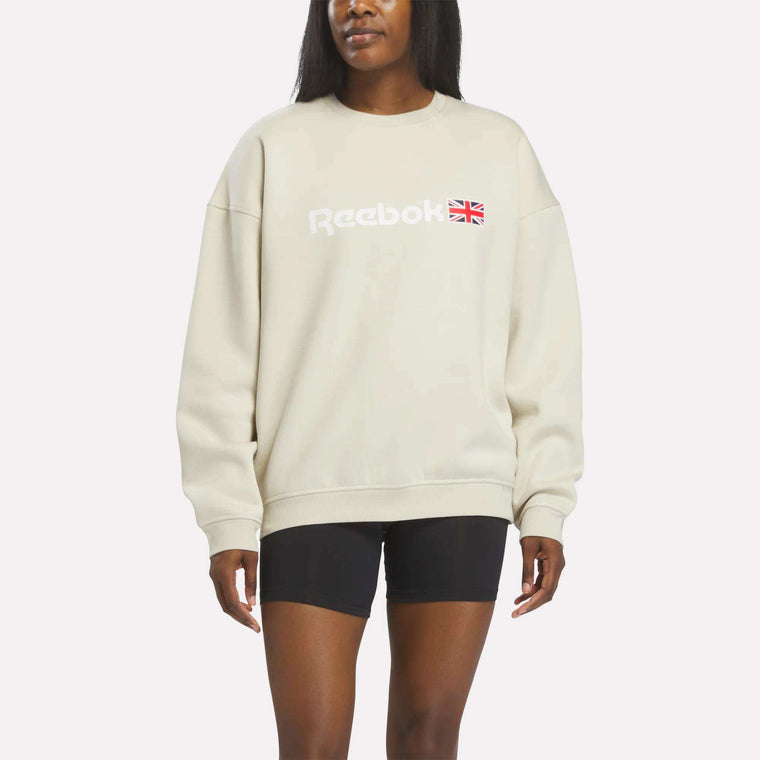 A woman wearing a Reebok Crewneck Sweatshirt in Linen and black shorts stands against a plain white background.