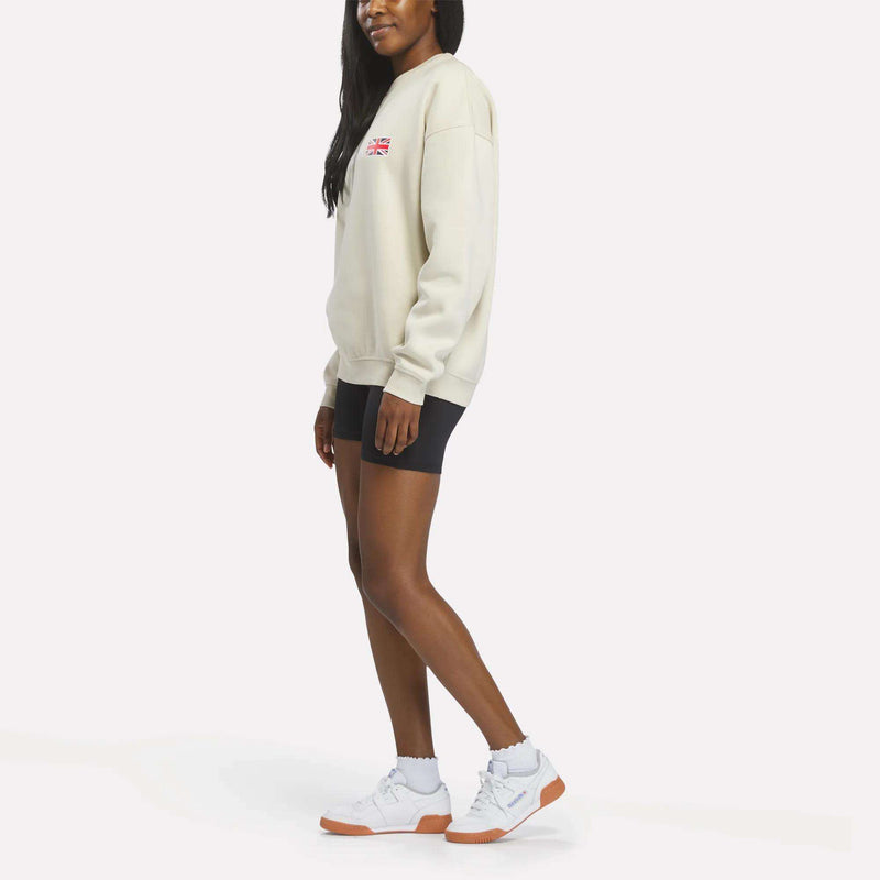 A woman stands against a plain light background wearing a relaxed fit Reebok Crewneck Sweatshirt in cream, featuring the brand’s Union Jack logo. She pairs it with black biker shorts and white sneakers with gum soles.