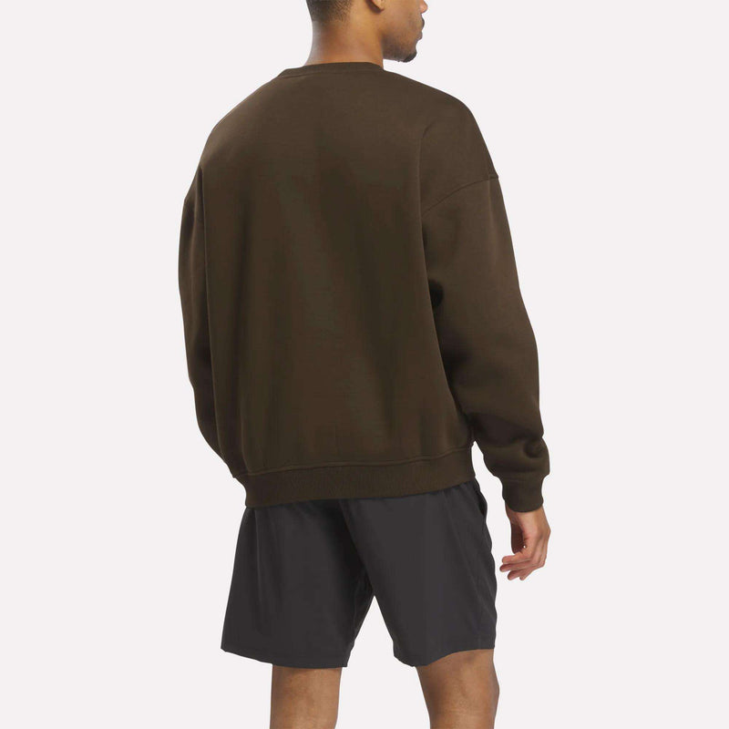 A man stands with his back to the camera, wearing a relaxed fit Crewneck Sweatshirt by Reebok in a deep brown shade featuring a subtle Union Jack logo, paired with black shorts against a plain light background.