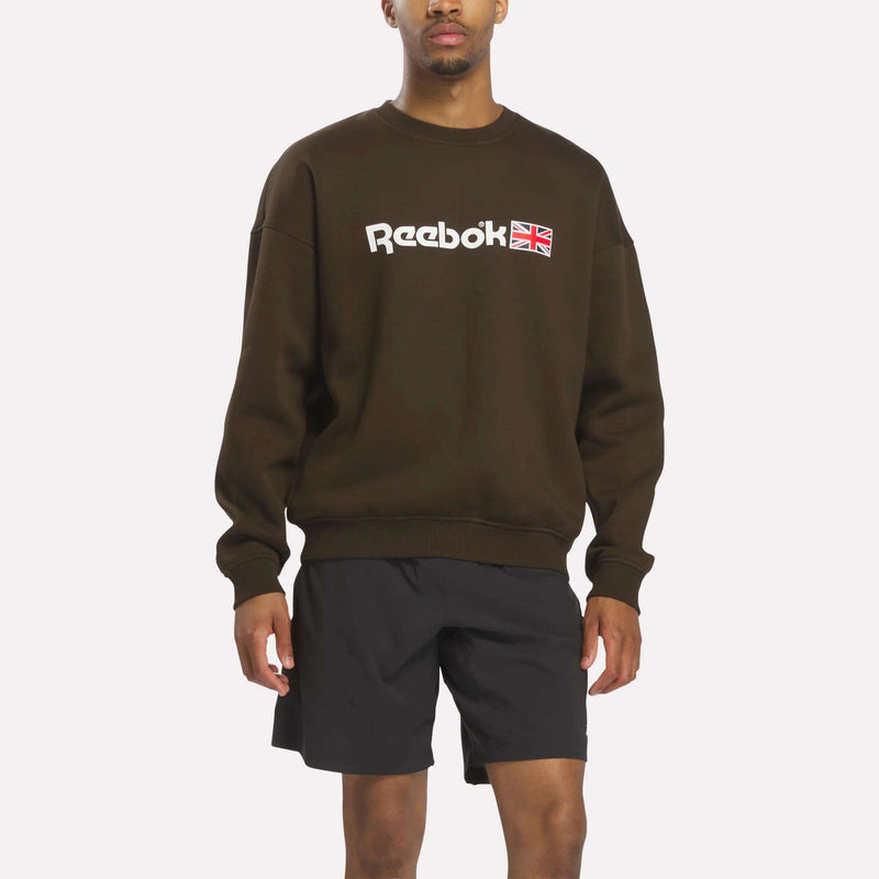 A man wearing a relaxed fit Reebok Crewneck Sweatshirt in espresso and black shorts stands against a plain white background. His face is partially out of frame.