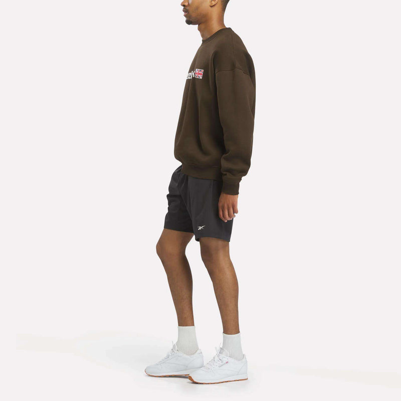 A person in a relaxed fit Reebok Crewneck Sweatshirt stands against a plain white background, facing sideways. The sweatshirt features the Union Jack logo and is paired with black shorts, white socks, and white sneakers.