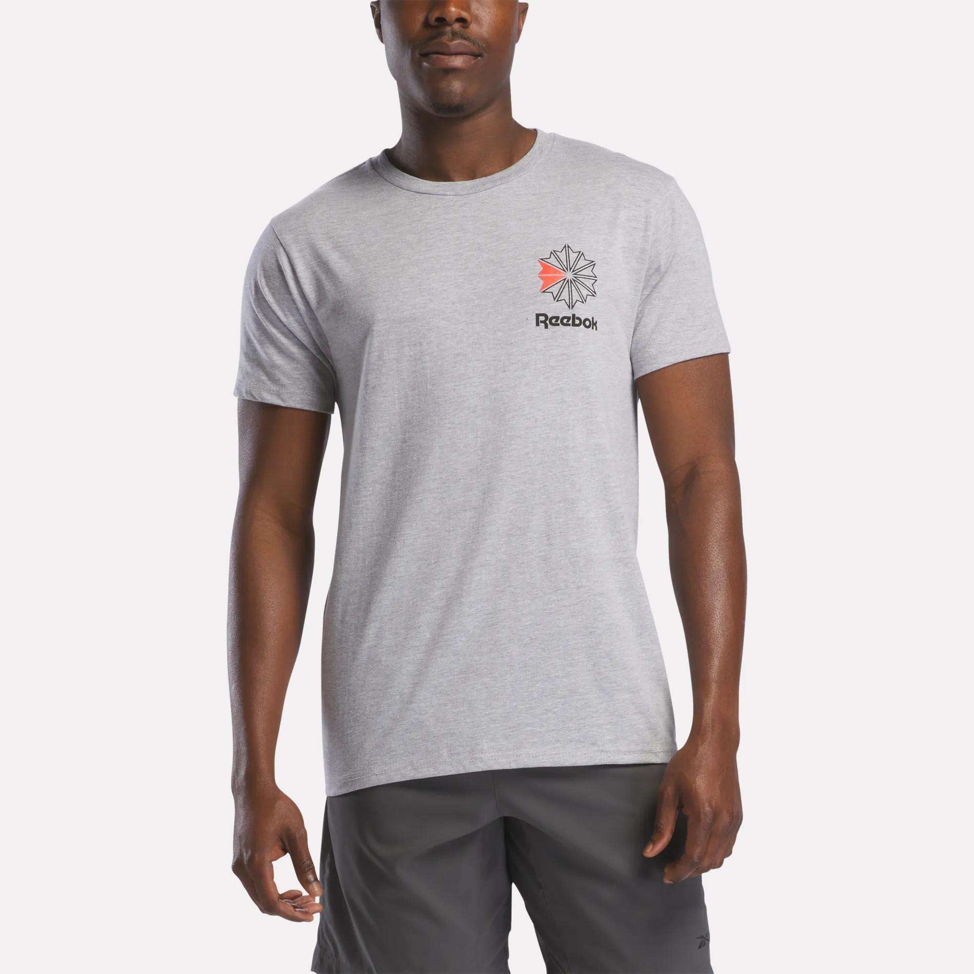 A man stands against a plain white background wearing a light gray Reebok Men's Starcrest Logo Graphic T-Shirt with a small logo on the left chest, paired with dark gray shorts.