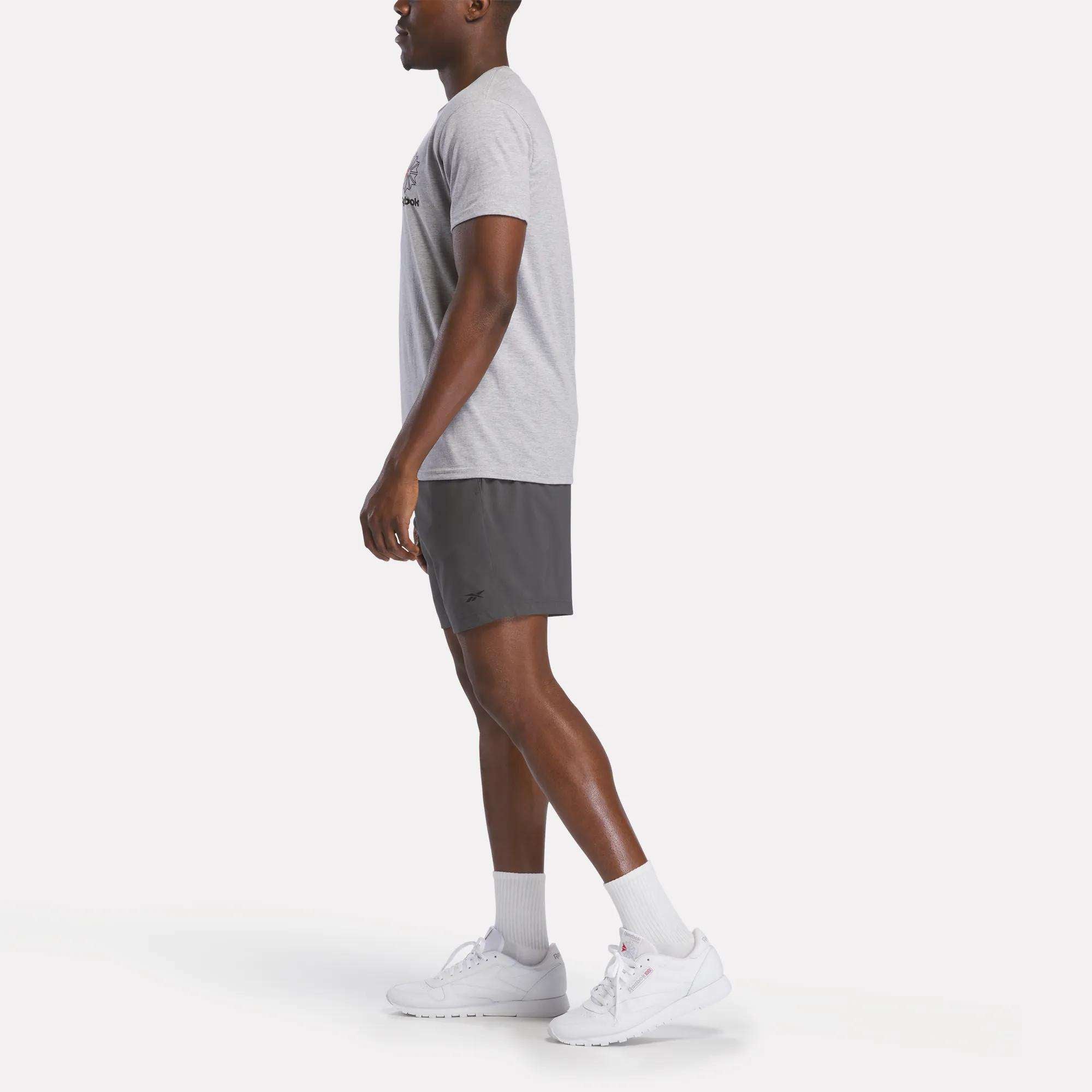 A man stands in profile against a white background wearing a light gray Reebok Men's Starcrest Logo Graphic T-Shirt, dark gray athletic shorts, white crew socks, and white Reebok sneakers.