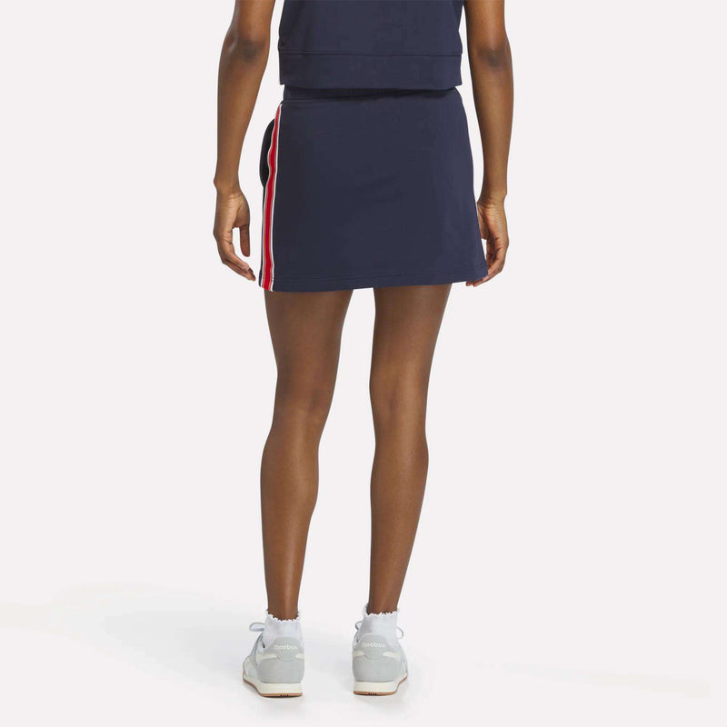 A woman faces away, wearing the Reebok Women's Logo Skirt in navy with red and white side stripes, paired with a matching navy top, white socks, and light sneakers against a plain white background.
