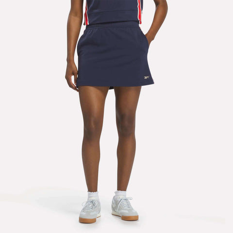 A person stands with one hand in the pocket of a Reebok Women's Logo Skirt in Vector Navy, paired with a matching top and white sneakers with light blue socks, set against a plain light background.