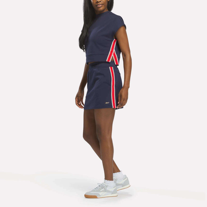 A woman wears the Reebok Women's Logo Skirt in navy blue with red and white side stripes, paired with a matching sleeveless top and light gray sneakers. She poses slightly turned to the side against a plain white background.