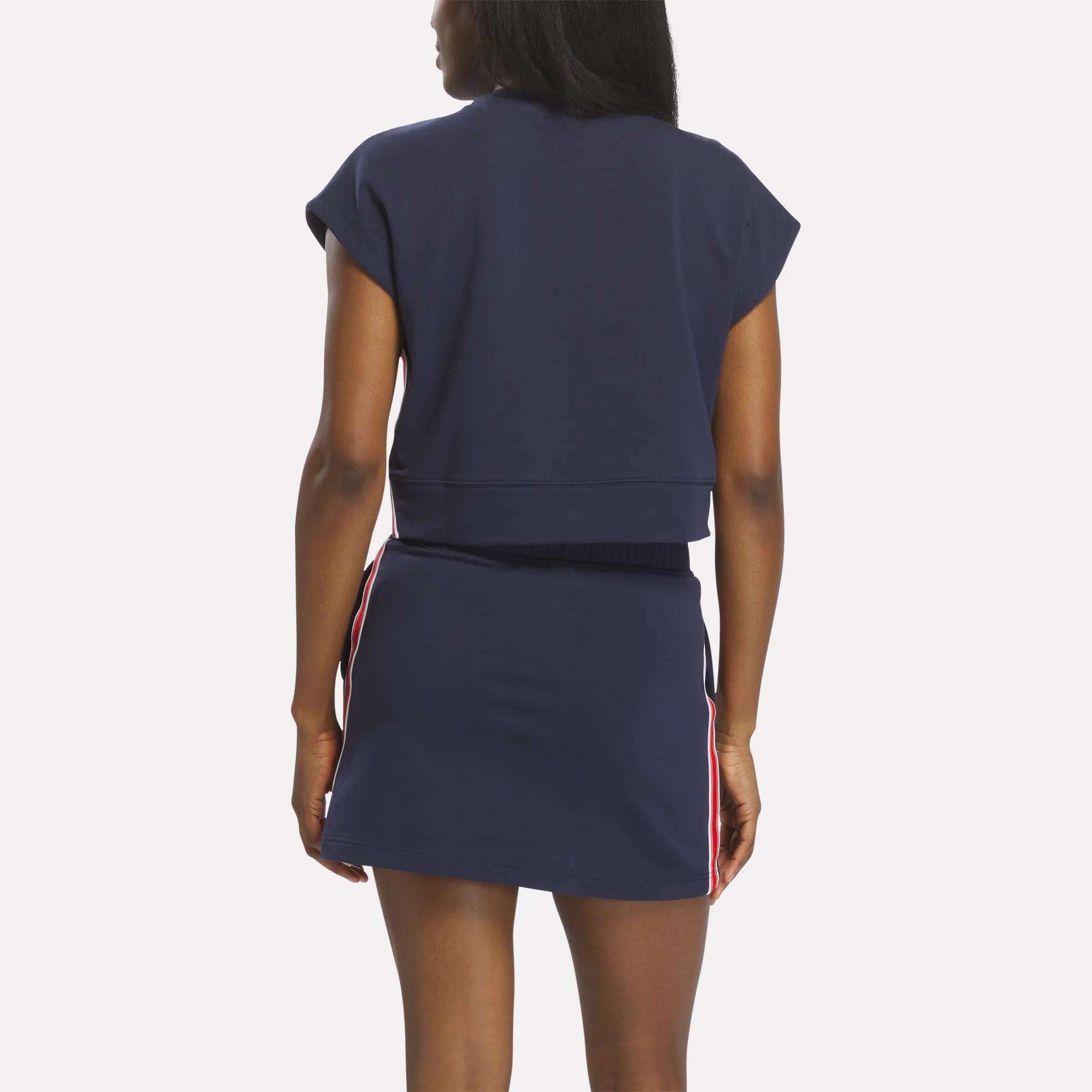 A woman stands facing away, wearing a navy blue Reebok Women's Logo Short Sleeve Crewneck Sweatshirt and matching mini skirt with red and white side stripes and a subtle gold chrome logo, set against a plain white background.