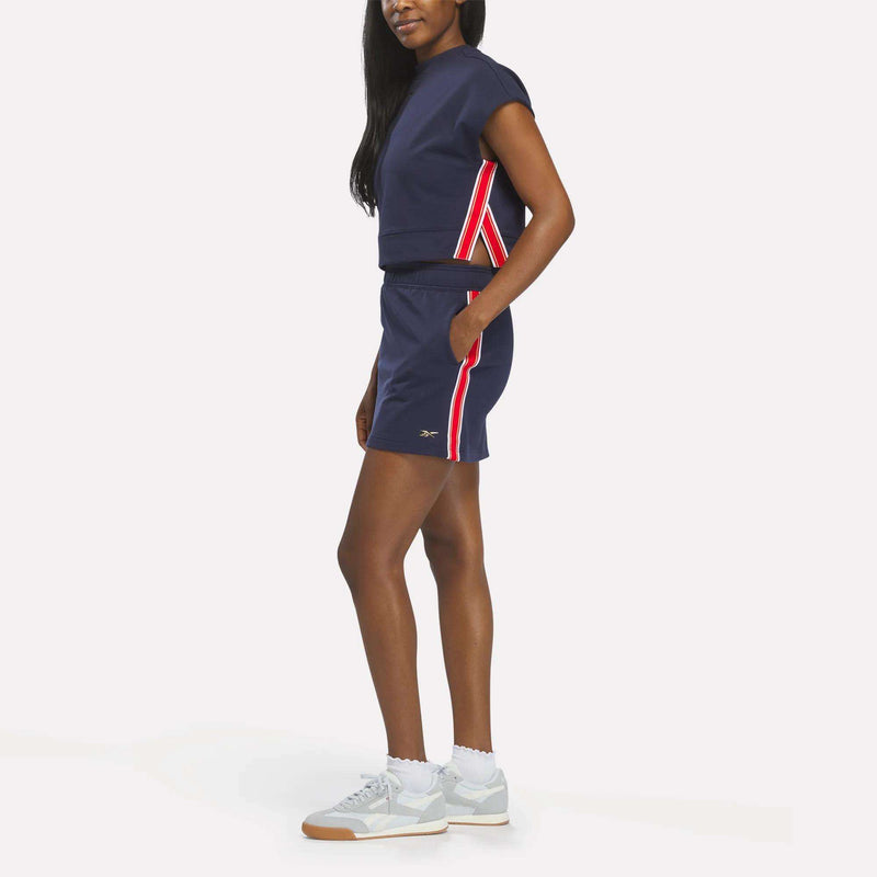 A woman stands sideways wearing a navy blue Reebok Women's Logo Short Sleeve Crewneck Sweatshirt with matching shorts featuring red and white striped accents, white socks, light gray Reebok sneakers, and a gold chrome logo on her top. Her hair is down.