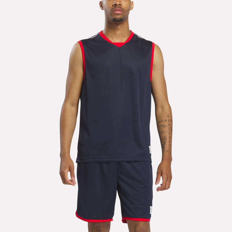 A man wears the Reebok Men's United Jersey Tank in Vector Navy, paired with matching shorts featuring red trim, standing against a plain light background.