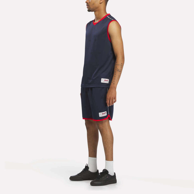 A man stands sideways wearing the Reebok Men's United Jersey Tank in navy blue with red trim, matching shorts, white socks, and black sneakers against a plain white background.