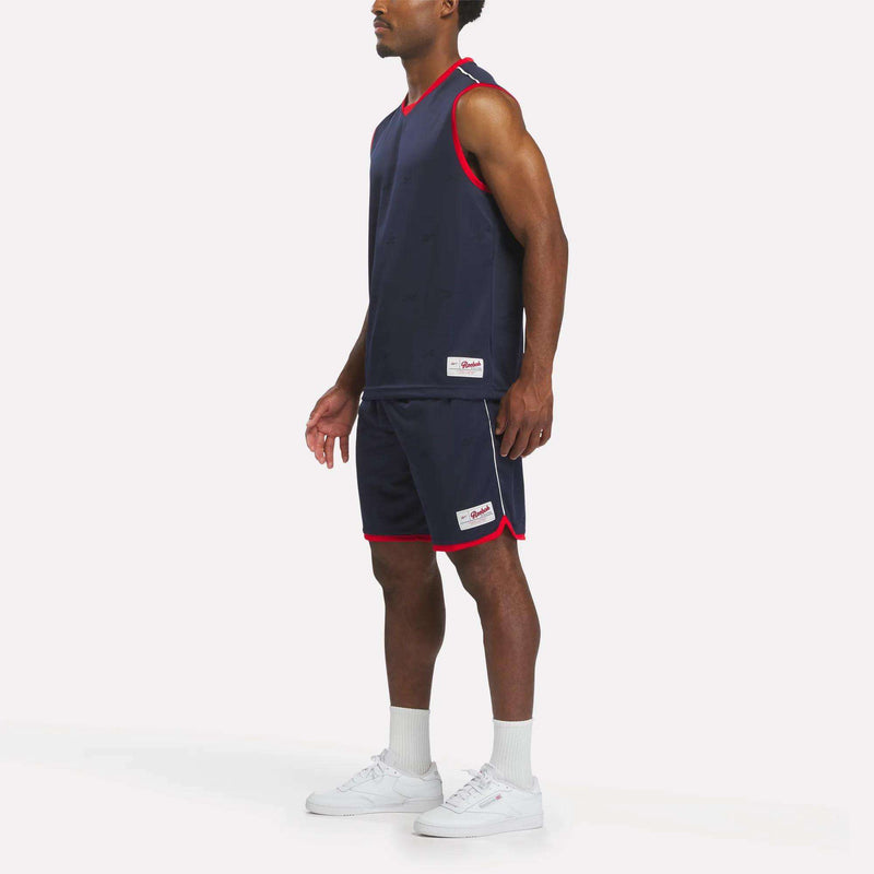 A person stands against a plain white background wearing the Reebok Men's United Jersey Tank in navy blue, paired with matching shorts featuring red trim, along with white socks and white sneakers for a classic soccer-inspired look.