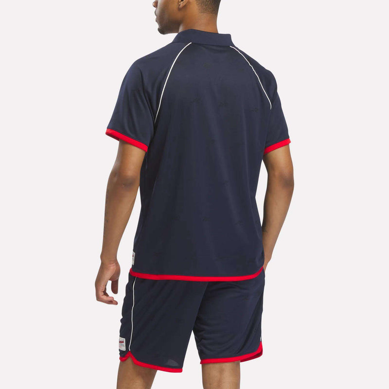 A person faces away, wearing a navy blue Reebok Men's United Jersey Polo Shirt and matching shorts with red trim and white piping, set against a plain white background.