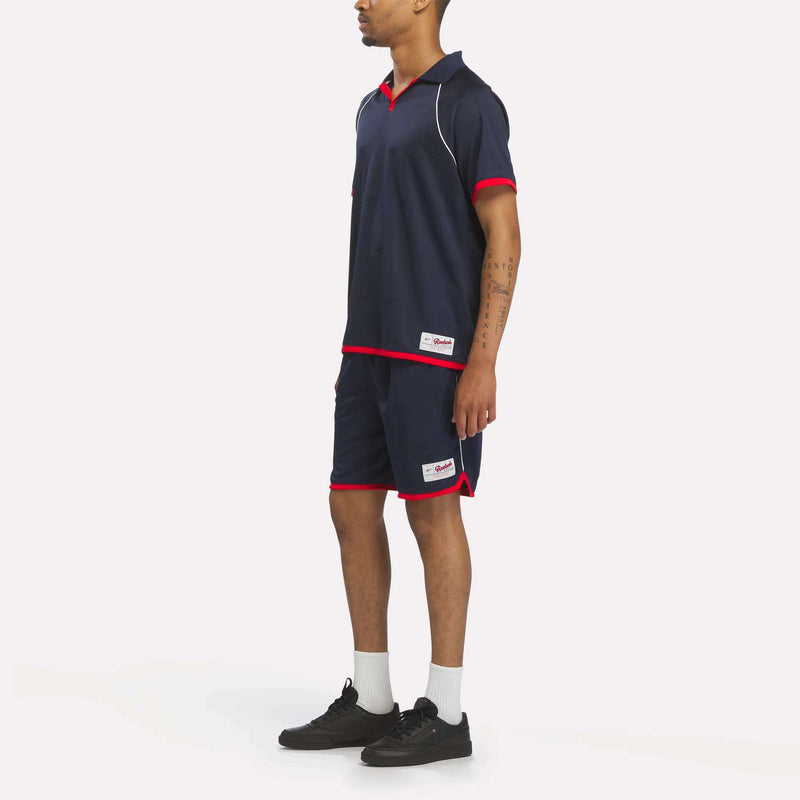 A man faces slightly sideways, wearing a Reebok Men's United Jersey Polo Shirt in navy blue with red trim, paired with matching shorts, white socks, and black sneakers against a white background.