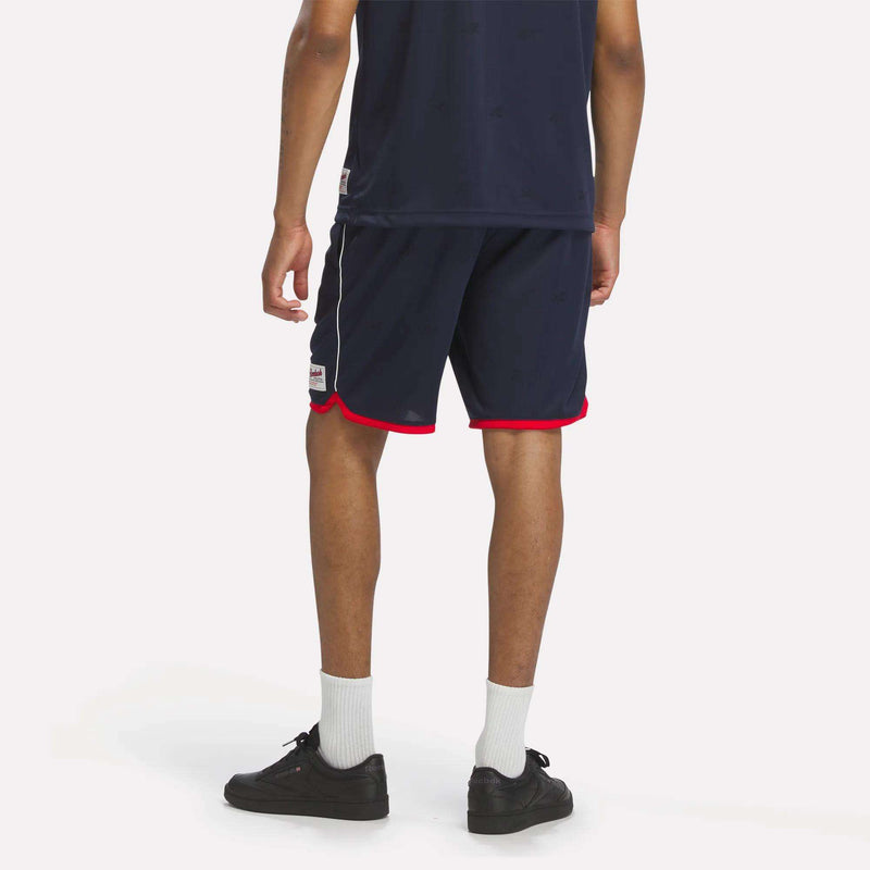 A person faces away, displaying a sporty look in a navy blue athletic shirt and Reebok Men's United Shorts 9" with red trim, paired with white crew socks and black sneakers against a plain light background.
