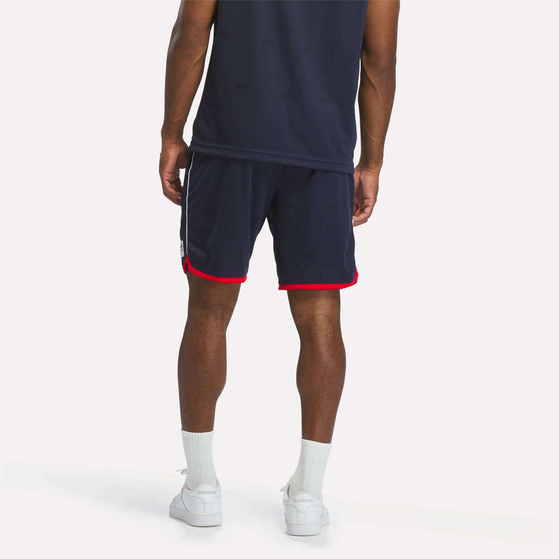 A person stands facing away, dressed in a navy shirt and Reebok Men's United Shorts 9" in navy with red trim, exuding a sporty style. The look is completed with white socks and white sneakers, set against a plain light background.
