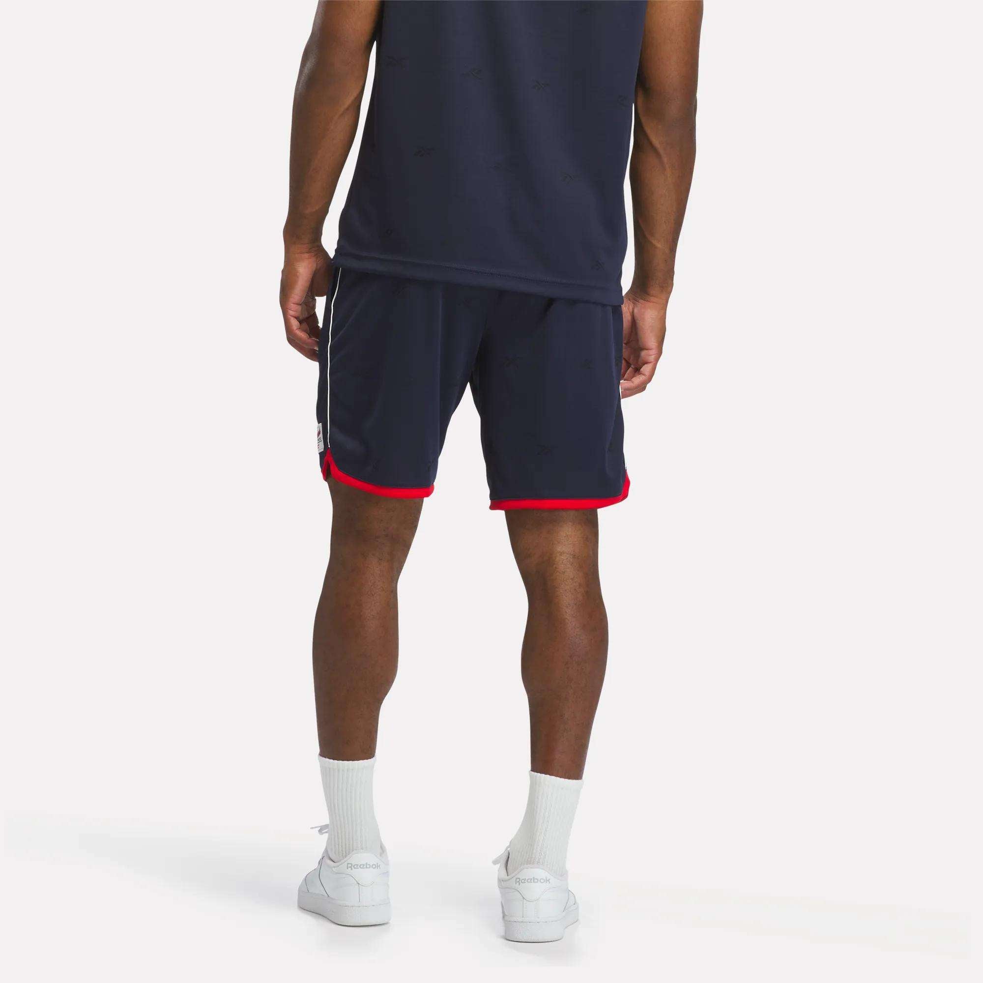 A person wearing Reebok Men's United Shorts 9" in navy with red trim, a matching shirt, white socks, and white sneakers stands with their back to the camera against a plain background.